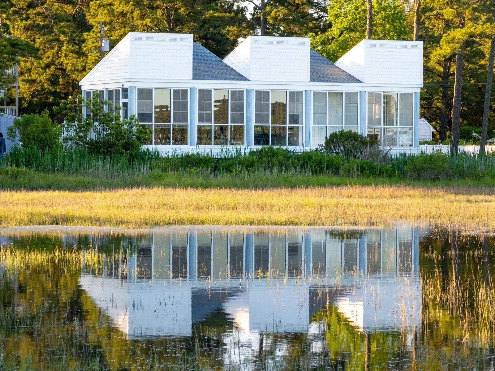 A two-story white house with many windows reflected in a body of water in the foreground